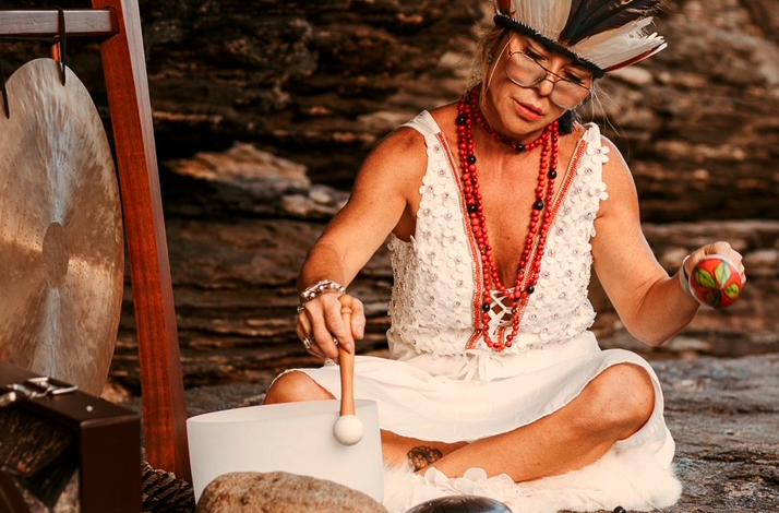 Individual performing a sound healing ritual with a bowl and gong on seaside rocks