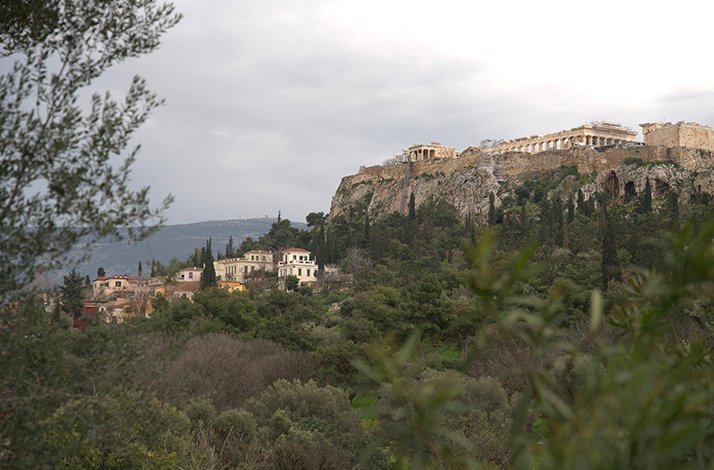 A view of the Acropolis of Athens and the Plaka neighborhood under a cloudy sky, framed by green trees.