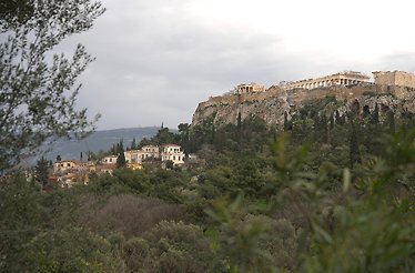 A view of the Acropolis of Athens and the Plaka neighborhood under a cloudy sky, framed by green trees.