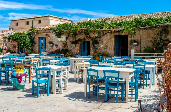 An outdoor cafe with white tables and blue chairs under a clear blue sky in Aegina, Greece..
