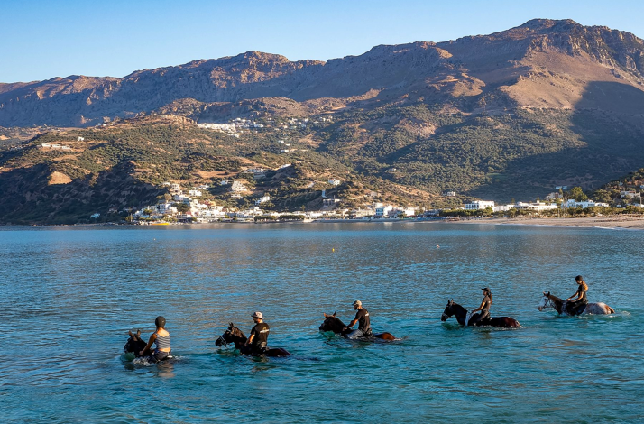 People riding horses in the sea at Palamida Cove, Hydra, with mountains and village in the background.