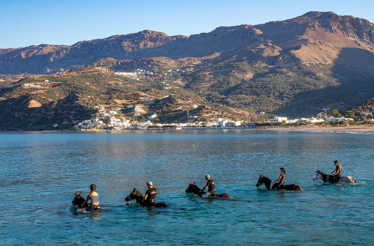 People riding horses in the sea at Palamida Cove, Hydra, with mountains and village in the background.