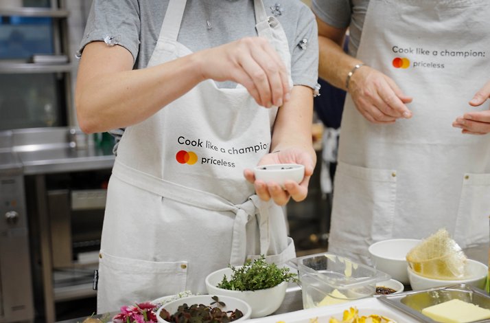 Hands sprinkle seasoning from a small white bowl over fresh herbs and ingredients, bringing creativity to life in a lively cooking class.