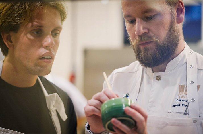 A guest watches closely as a chef demonstrates a delicate technique, sharing expertise and inspiration in the kitchen.