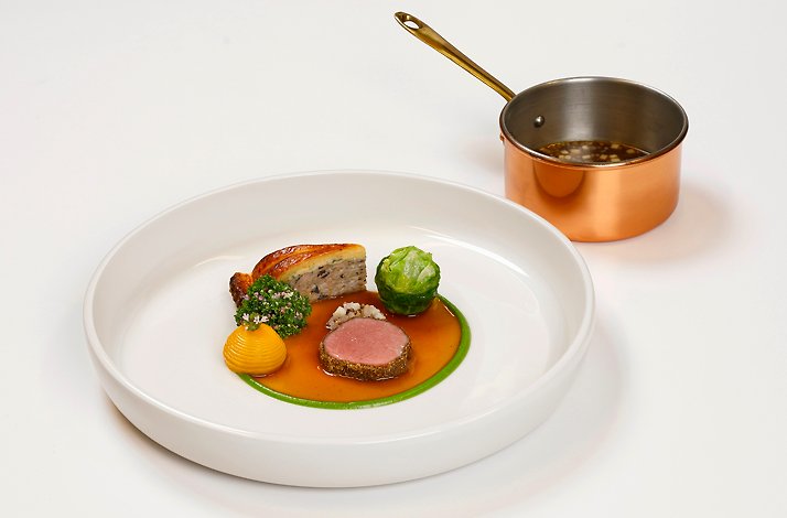 A refined main course artfully arranged on a wide porcelain plate, accompanied by a copper saucepan, creating a sense of classical technique and warm, professional kitchen elegance.