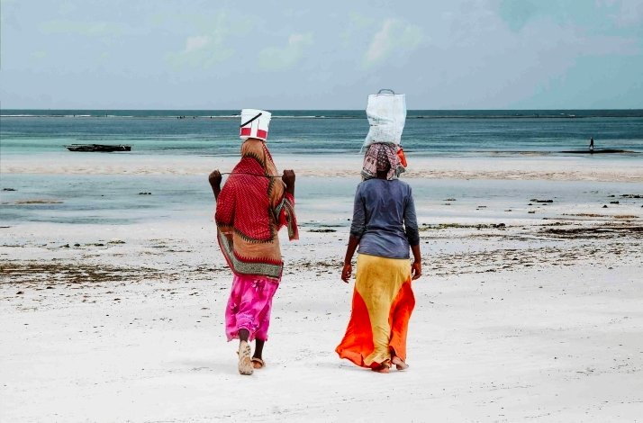 Two women walking on the beach, balancing water bottles on their heads, enjoying a sunny day by the ocean.