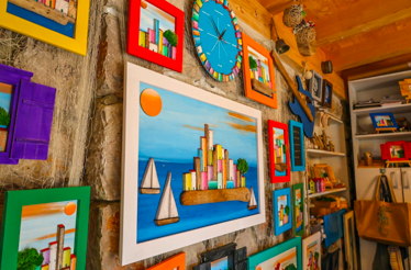 Colorful framed artworks of seaside towns and boats, made from driftwood and painted elements.