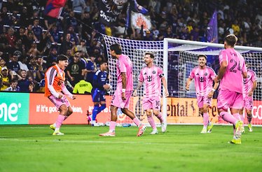 Inter Miami CF players in pink uniforms gather near the goal during a match, with fans in the background.