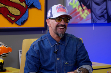YouTuber Yiğit Aydın wearing a denim jacket, cap, and tinted glasses sits in a gallery space decorated with pop-art works and collectible figures
