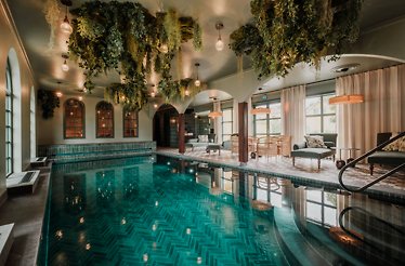 The serene indoor spa pool at Lejondals Slott in Bro, Sweden, glows beneath hanging greenery and warm lights, creating a tranquil retreat. © Countryside Hotels