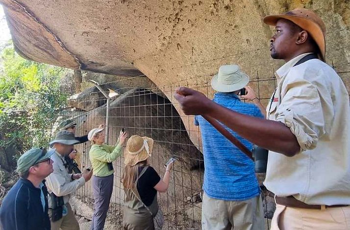 A group of people gathered around a large rock formation, with a man wearing a hat prominently visible among them.