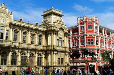 Ornate historic buildings with people walking in front, under a bright blue sky.