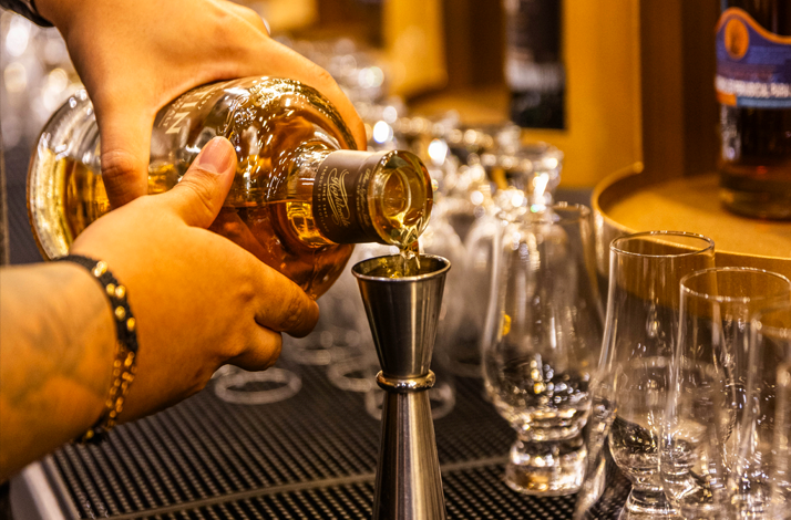 Hands pouring whisky from a bottle into a jigger over a bar with aligned glasses
