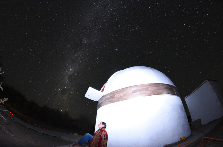 Person observing the night sky next to a white dome with the Milky Way visible above the horizon