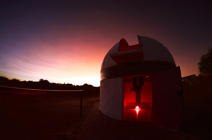Open observatory dome with red light inside under a colorful sky at dusk