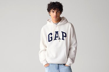 A man wearing a white GAP hoodie featuring a bold blue logo across the chest. The hoodie has a relaxed fit, with a drawstring hood and ribbed cuffs, giving it a casual and comfortable look