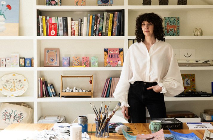 Brianda Fitz-James Stuart posed in her Madrid art studio, wearing a white blouse, with art books and painted canvases displayed on shelves. 