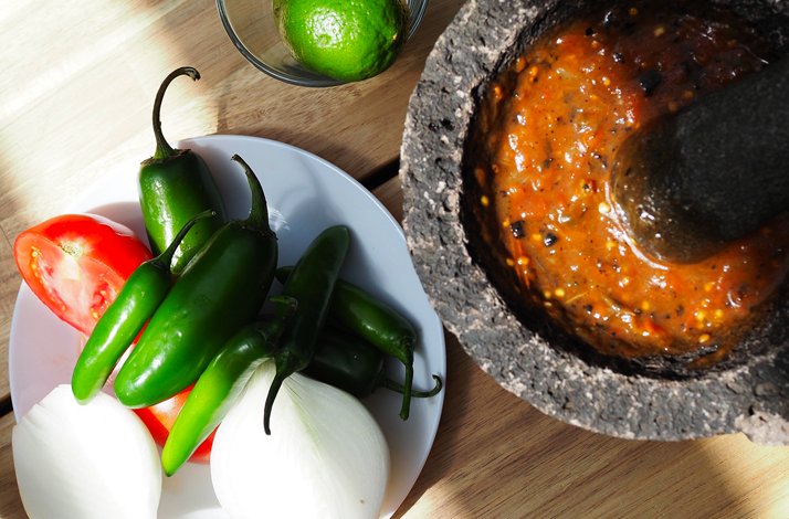 Fresh ingredients including chiles, tomatoes, onion, and lime arranged next to a molcajete for a traditional salsa-making ritual.