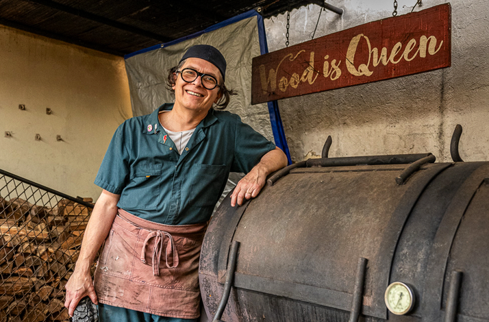 Chef Mario Rocero standing beside a wood smoker with a 'Wood is Queen'