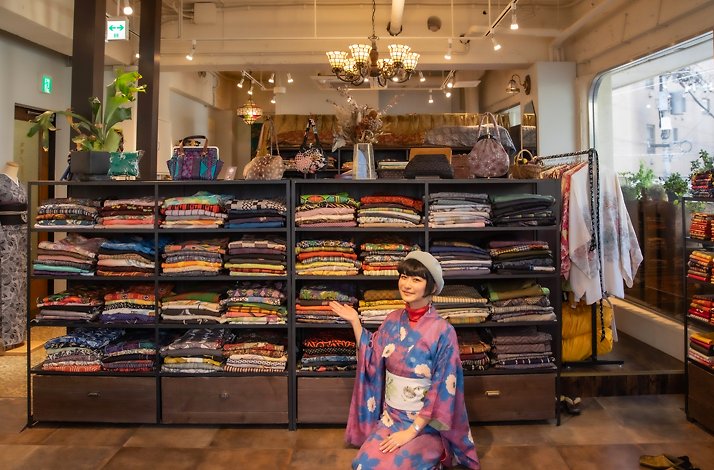 Sankak sitting next to shelves with kimonos while wearing a kimono herself.