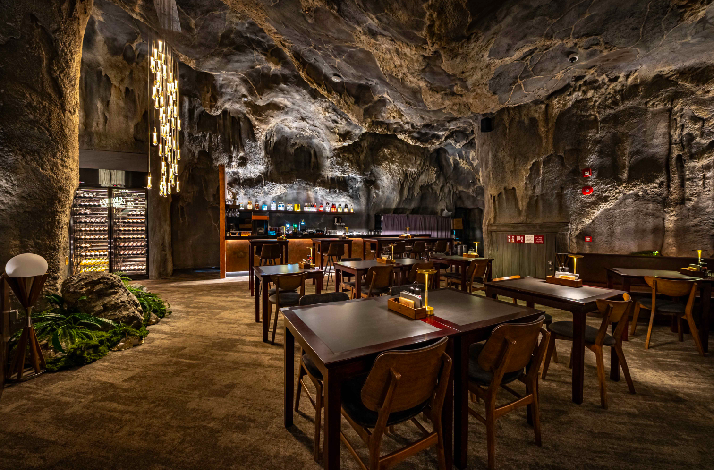 The interior of The Cavern restaurant at Mandai Wildlife Reserve, featuring realistic rock-hewn walls, dark wood furniture, and warm lighting.