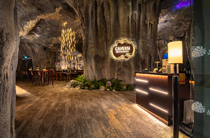 Entrance of The Cavern restaurant, Mandai Wildlife Reserve, featuring a backlit logo on a rock wall, wood reception desk, and cave-like interior.