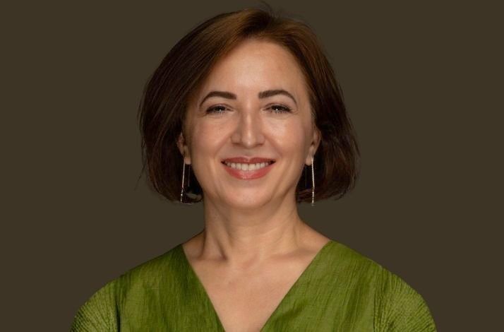 A studio portrait of Bahriye Çeri smiling against a dark brown background. She has short brown hair, long silver vertical earrings, and is wearing a V-neck, pleated forest green blouse. The shot is framed from the shoulders up.