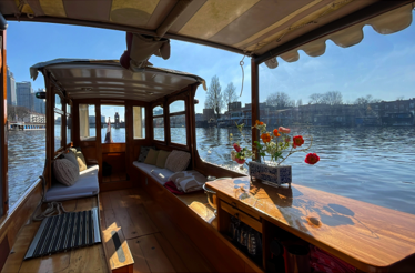 Cozy wooden boat interior with flowers, cushions, and canal views under sunny Amsterdam skies.