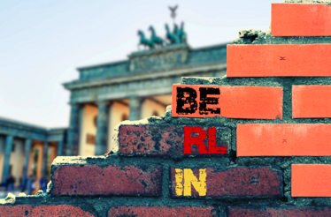 A partially broken brick wall with BERLIN painted on it, with the Brandenburg Gate in the background.