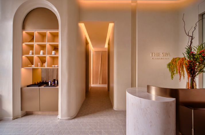 Elegant, minimalist spa reception area featuring warm beige tones, an arched display shelf with neatly arranged products, a curved marble counter, and a softly lit hallway leading to treatment rooms.
