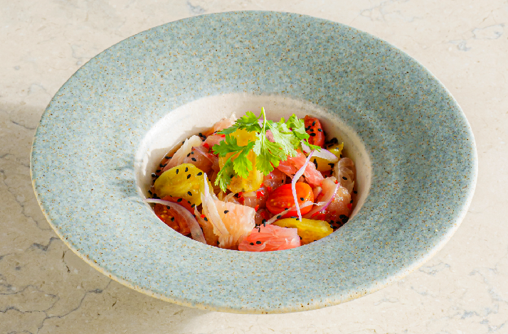 A refined ceviche-style dish served in a wide-rimmed speckled blue ceramic bowl, featuring colorful heirloom tomatoes, delicate fish, pickled red onion, fresh cilantro, and black sesame seeds, presented at Rockfish's cliffside restaurant in Uluwatu.