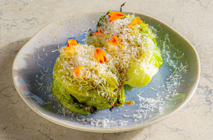 An elegant plated dish at Rockfish featuring two charred baby gem lettuce heads generously topped with finely grated white cheese and garnished with vibrant orange edible flower petals, served on a speckled blue-grey ceramic plate against a marble surface.