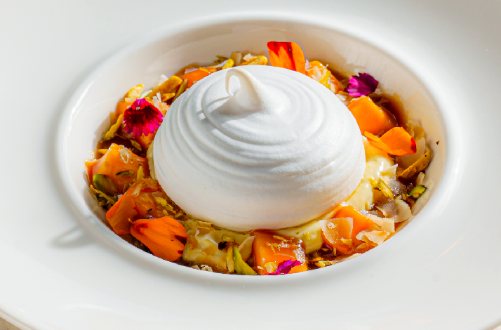 A stunning dessert at Rockfish featuring a perfectly swirled dome of white meringue or cream atop a bed of tropical fruit, crushed pistachios, coconut flakes, and vibrant edible flowers in pink and orange, served in a white ceramic bowl.