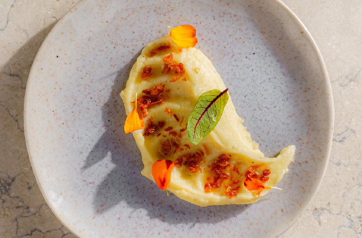 A delicately crafted half-moon shaped pasta or dumpling, glazed in a golden butter sauce and adorned with crispy shallots, a small burgundy leaf, and bright orange nasturtium petals, plated artfully on a speckled ceramic dish at Rockfish.