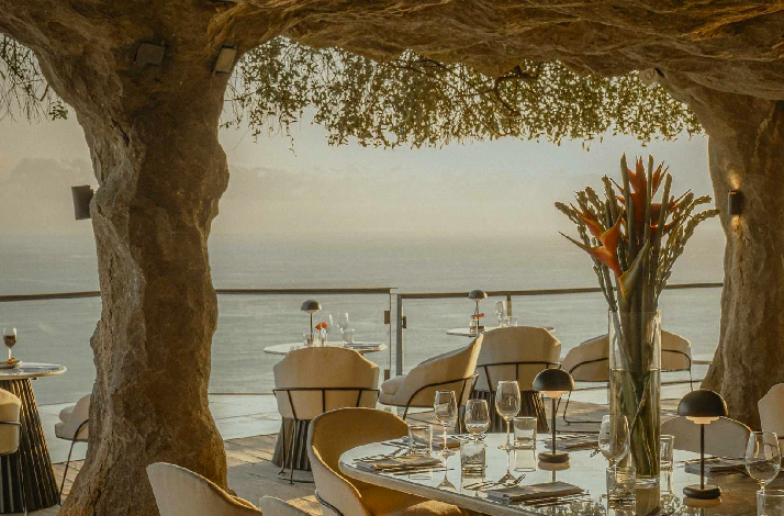 An elegant cliffside dining setup inside a cave-like structure with natural stone pillars and vine-covered ceiling. Tables are set with glassware, place settings, and bird-of-paradise flower arrangements, with plush cream-colored chairs. The open side reveals a panoramic ocean view in soft golden light.