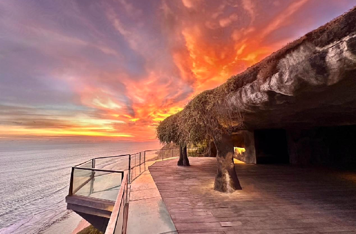 A dramatic sunset view from a cliffside cave walkway with a wooden deck and metal railing overlooking the ocean. The sky is ablaze with vivid orange, pink, and purple clouds. Tree trunk-like stone pillars support a natural rock overhang draped with greenery, with warm lighting illuminating the cave entrance.