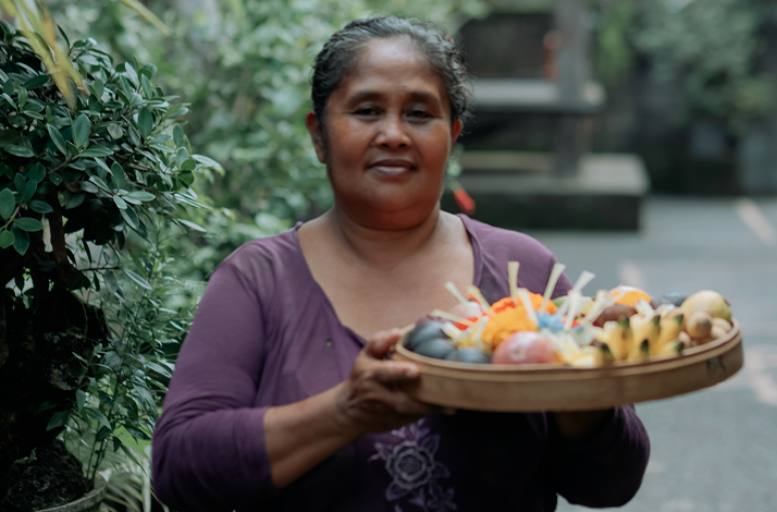 Balinese woman holding a tray of traditional food in a family compound.
