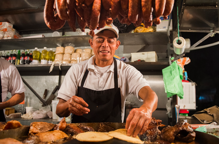 Street cook preparing tacos on a hot grill surrounded by hanging meats.