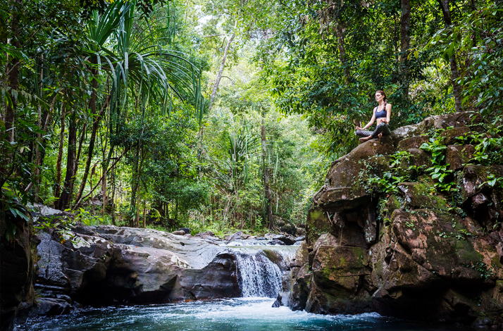 Woman meditating on a rocky cliff beside a waterfall in the lush rainforest of The Datai Langkawi.