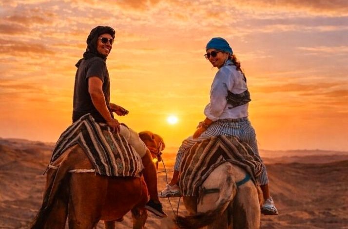 Two people riding camels in the Agafay Desert at sunset, with warm orange skies and vast sandy terrain in the background.