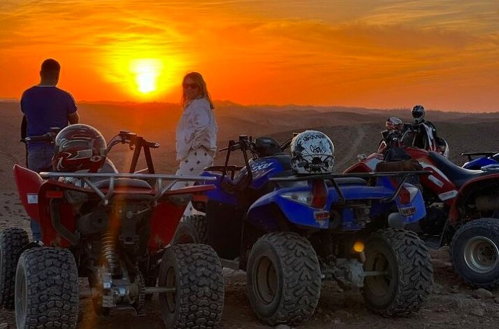 Group of people standing beside quad bikes in the desert, watching the sun set over rugged, open landscape.