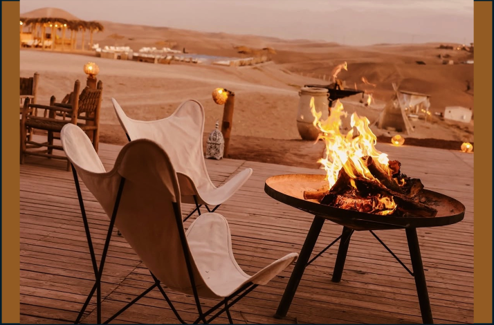 Cozy campfire burning in a fire pit on a wooden deck, with empty lounge chairs overlooking the sandy desert and distant tents.