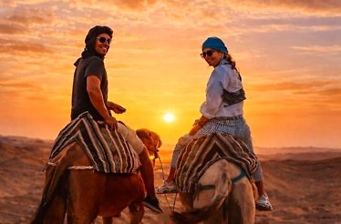 Two people riding camels in the Agafay Desert at sunset, with warm orange skies and vast sandy terrain in the background.