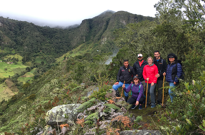 Group of hikers standing at a mountain viewpoint surrounded by Andean vegetation, with sweeping views of valleys and cloud-covered peaks in the background.