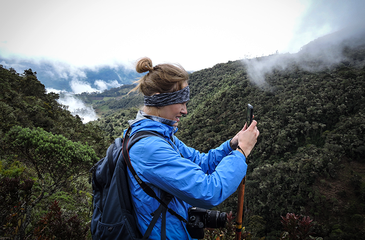 Hiker in a blue jacket and backpack using a smartphone to photograph a misty mountain landscape overlooking a cloud forest in the Andes.