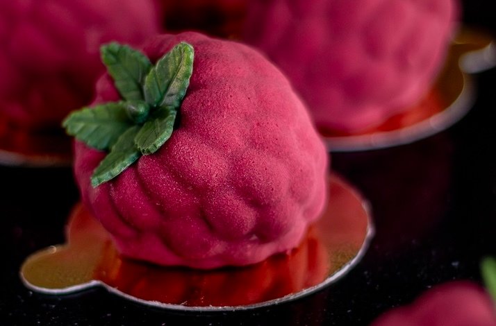 Realistic raspberry-shaped mousse dessert with green leaves on a gold base.