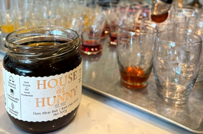 A close-up of a 'House of Hunny' raw oak honey jar in the foreground, with several small glass cups filled with different types of honey arranged on a metal tray for tasting in the blurred background.
