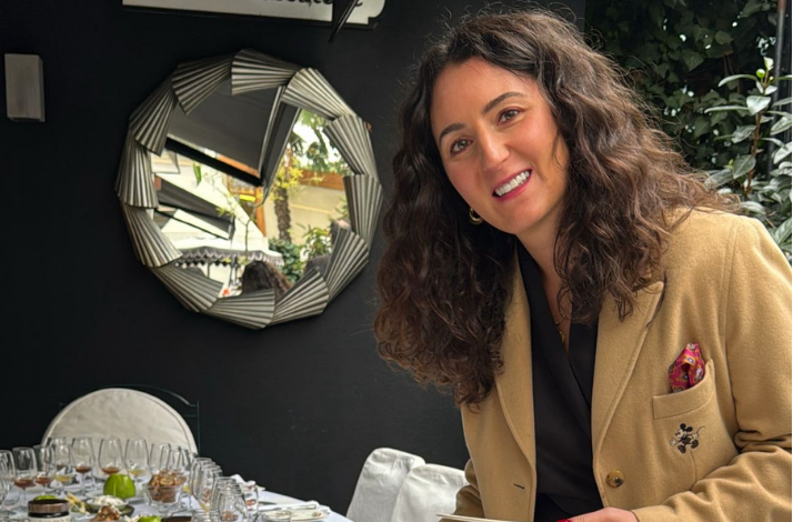 Chef Elif Yalın, wearing a tan blazer with a Mickey Mouse embroidery, smiles next to a honey tasting table. A decorative sunburst mirror is visible on the dark wall behind her.