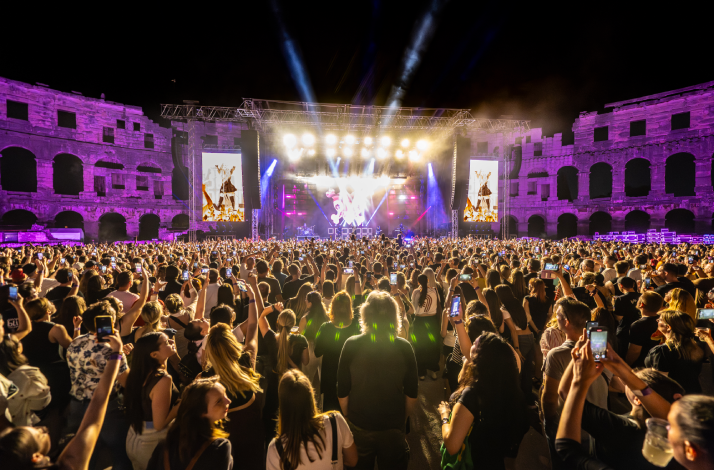 A vibrant night concert scene inside the Pula Arena in Pula, Croatia, showing a dense crowd of people with their phones raised.