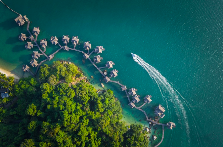 Aerial view of overwater villas, green island, and a boat on turquoise waters at Pangkor Laut Resort.